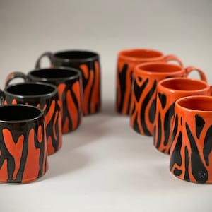 May include: A collection of ceramic mugs with a tiger stripe design. The mugs are orange with black stripes and black interiors. Each mug has a handle and a glossy finish. The mugs are arranged in a semi-circle.