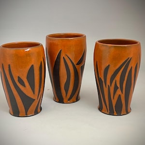 May include: Three ceramic vases with a vibrant orange glaze and dark brown, abstract leaf-like patterns. The vases have a smooth, glossy finish and a cylindrical shape, perfect for displaying flowers or as decorative pieces.