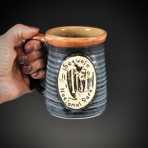 May include: A gray ceramic mug with a brown rim and a white oval logo that says "Sequoia National Park". The logo features a stylized image of a giant sequoia tree.