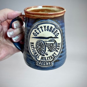 May include: A blue and brown ceramic mug with a white design featuring the words "Gettysburg National Military Park 1863" and a cannon.