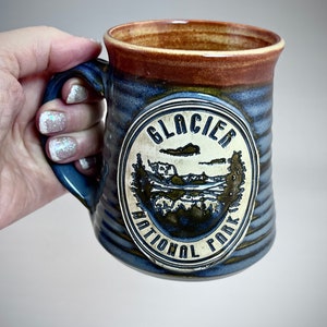 Puede incluir: Taza de cerámica con un esmalte azul y marrón, con un emblema del Parque Nacional Glacier. La taza tiene un asa resistente y un exterior texturizado. El emblema representa un paisaje con las palabras "Glacier National Park".