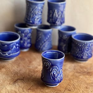 May include: A collection of eight ceramic cups in varying shades of blue, each featuring unique raised white relief patterns. The cups are cylindrical with a slightly flared rim and a small footed base, showcasing distinct hand-applied decorative motifs.