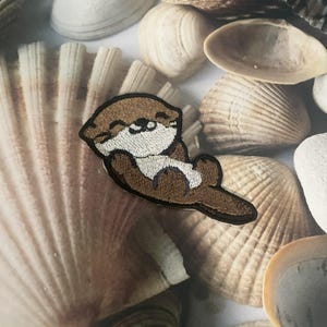 May include: A brown and white embroidered otter patch, lying on a bed of seashells.