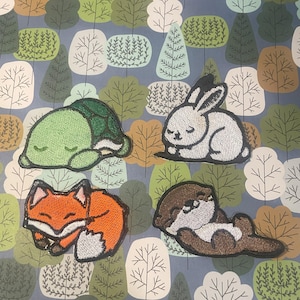 May include: Four embroidered patches featuring sleeping animals: a green turtle, a white rabbit, an orange fox, and a brown otter. The patches are on a blue background with a repeating pattern of green trees.