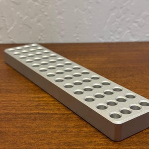 May include: A silver rectangular object with numerous evenly spaced circular holes. The object is made of metal and sits on a wooden surface. The design suggests a tool or organizational item.