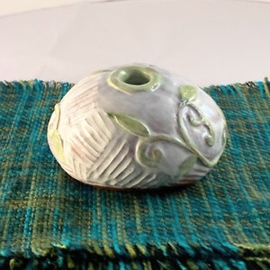 May include: A small, white ceramic vase with a green floral design. The vase is shaped like a rounded stone and has a hole in the top for flowers.