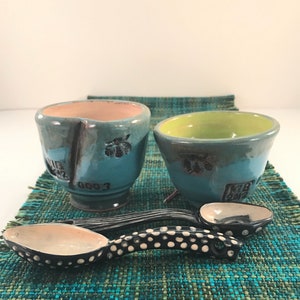 May include: Two small blue ceramic bowls with a green and pink glaze, and two black and white speckled ceramic spoons on a green and blue woven placemat.