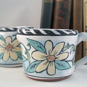 May include: Two white ceramic mugs with black trim and floral designs. The mugs have a blue and green floral pattern with yellow flowers. The mugs are sitting on a white surface.
