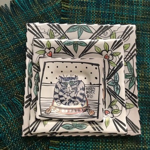 May include: Three white ceramic plates with black and green floral designs. The smallest plate features a black and white illustration of a cat sitting on a red and white cushion. The signature "Karen Baker 9/15" is visible on the plate.