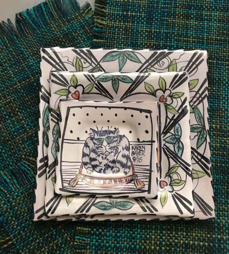 May include: Three white ceramic plates with black and green floral designs. The smallest plate features a black and white illustration of a cat sitting on a red and white cushion. The signature "Karen Baker 9/15" is visible on the plate.