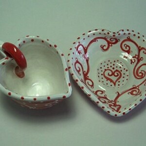 Puede incluir: Un juego de taza y platillo de cer&aacute;mica en forma de coraz&oacute;n, blanco con lunares rojos y un dise&ntilde;o de espiral rojo. El platillo tiene un coraz&oacute;n rojo en el centro.