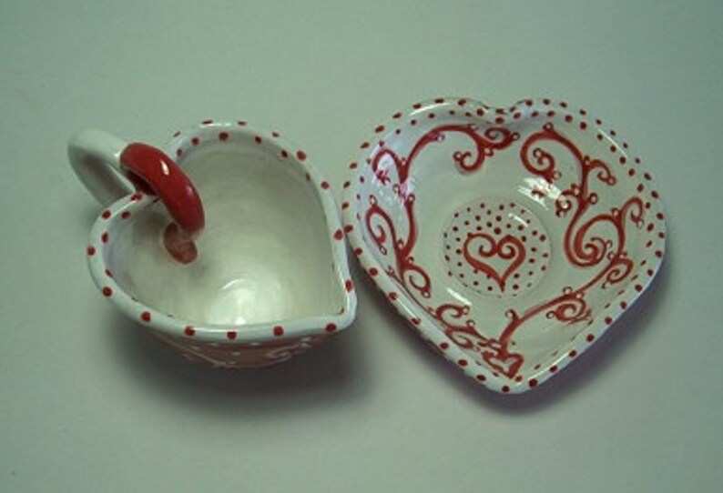 Puede incluir: Un juego de taza y platillo de cer&aacute;mica en forma de coraz&oacute;n, blanco con lunares rojos y un dise&ntilde;o de espiral rojo. El platillo tiene un coraz&oacute;n rojo en el centro.