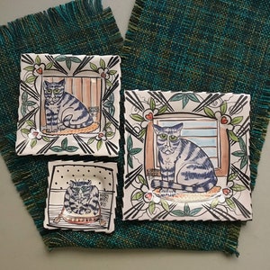 May include: Three ceramic square plates with hand-painted designs of cats. Each plate features a different cat with a unique expression and pose. The plates are decorated with black and white lines, green leaves, and red hearts. The text "KAREN BAKER" is visible on each plate.