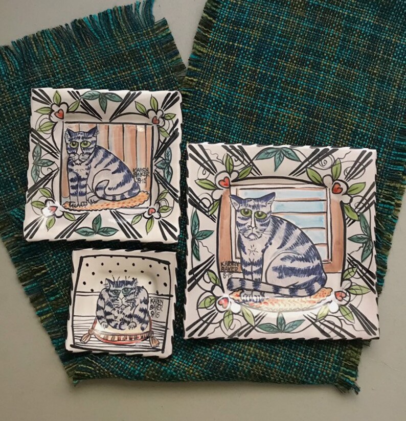 May include: Three ceramic square plates with hand-painted designs of cats. Each plate features a different cat with a unique expression and pose. The plates are decorated with black and white lines, green leaves, and red hearts. The text "KAREN BAKER" is visible on each plate.