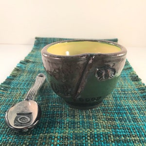 May include: A small, green ceramic bowl with a yellow interior and a brown glaze. The bowl has a unique, asymmetrical shape and a small, raised design on the side. The bowl is sitting on a green and blue woven placemat.