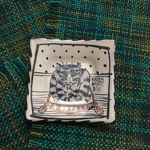 May include: White ceramic dish with a black and white illustration of a cat sitting on a pillow. The cat has blue eyes and a striped pattern. The dish is signed "Karen Baker 9/15".