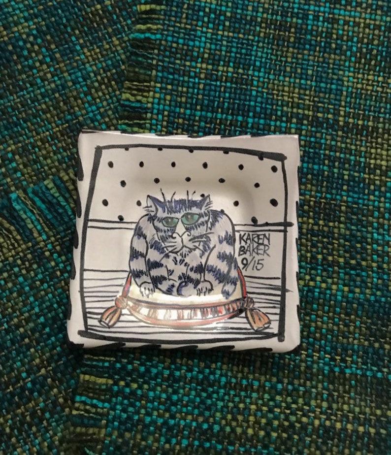 May include: White ceramic dish with a black and white illustration of a cat sitting on a pillow. The cat has blue eyes and a striped pattern. The dish is signed "Karen Baker 9/15".