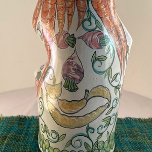 May include: White ceramic vase with a hand-painted design of carrots, peas, and turnips. The vase has a unique, slightly misshapen form.