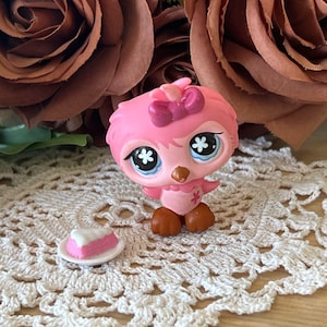 May include: Pink and brown plastic toy bird with a pink bow and blue eyes. The bird is standing on a white crocheted doily with a pink and white cake slice on a white plate.