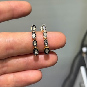 Puede incluir: Un par de pendientes de oro, cada uno con cuatro piedras ovaladas de color gris translúcido dispuestas en una fila vertical. Las piedras están engastadas en metal oscuro, creando un diseño lineal que sigue la curva de la estructura.