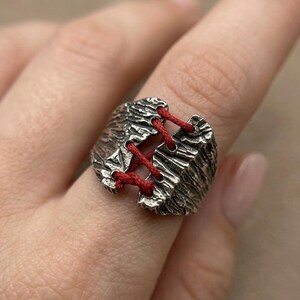Puede incluir: Un anillo plateado con un diseño texturizado y rugoso. El anillo presenta un diseño dividido, atado con un cordón rojo brillante. El anillo se lleva en un dedo, mostrando los intrincados detalles y el estilo único de la joya.