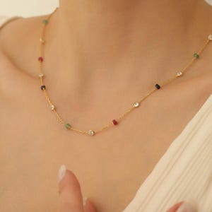 Puede incluir: Un collar de cadena dorada fina que presenta una serie de pequeñas piedras redondas y transparentes engastadas, intercaladas con diminutas cuentas de piedras preciosas en bruto de colores rojo, verde y negro, creando una joya refinada.