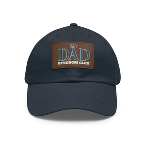 May include: A navy blue baseball cap with a curved peak. It has a brown patch displaying the words "DAD KINGDOM CLUB" in white and teal, and a small crown graphic.