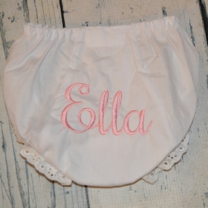 May include: White cotton baby bloomers with a pink embroidered name "Ella" and white lace trim.