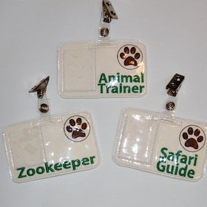 May include: Three white ID badge holders with clear plastic windows and brown paw print designs. The text on the badges reads "Animal Trainer", "Zookeeper", and "Safari Guide".