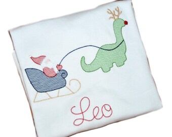 May include: White Christmas-themed t-shirt with an embroidered design of a Santa Claus in a sleigh being pulled by a green dinosaur with reindeer antlers. The name "Leo" is embroidered below the design.