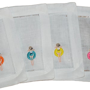 May include: Set of four white linen coasters with embroidered images of a woman in a swimsuit and a swim ring. Each coaster features a different color swim ring: orange, teal, yellow, and pink. The coasters have a stitched border.