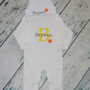May include: White baby onesie with long sleeves and snaps at the bottom. The onesie has a yellow letter "B" and the name "Bryson" embroidered on the front. A small orange basketball is embroidered next to the name. A white hat with the name "Bryson" and a small orange basketball embroidered on it is on top of the onesie.