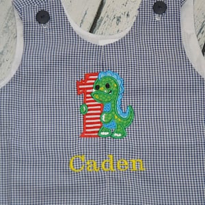 May include: Blue and white gingham baby overalls with a red and white number one and a green dinosaur applique. The name "Caden" is embroidered in yellow below the dinosaur.