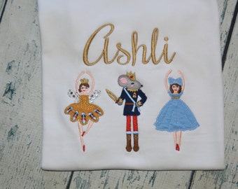 May include: White t-shirt with the name "Ashli" embroidered in gold thread. The shirt features three embroidered figures: a ballerina in a gold dress, a nutcracker in a blue and red uniform, and a ballerina in a blue dress.