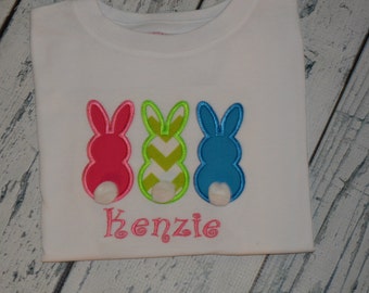 May include: White t-shirt with three embroidered bunny silhouettes in pink, green, and blue. The bunnies have white pom-poms for tails. The name "Kenzie" is embroidered below the bunnies in pink.