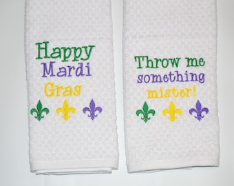 Puede incluir: Dos toallas de cocina blancas con texto bordado con temática de Mardi Gras. La primera toalla dice "Happy Mardi Gras" con tres flores de lis verdes, amarillas y moradas. La segunda toalla dice "Throw me something mister!" con tres flores de lis verdes, amarillas y moradas.