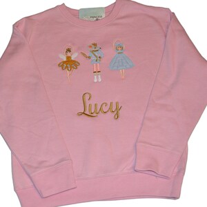 May include: Pink children's sweatshirt with a Nutcracker, ballerina, and mouse embroidered on the front. The name "Lucy" is embroidered in gold below the figures.