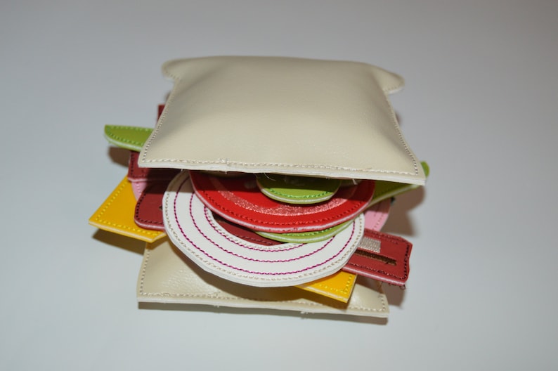 May include: A faux leather sandwich purse with a white top bun, red tomato, green lettuce, white onion, pink ham, and yellow cheese.