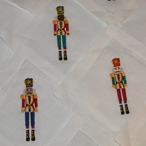 May include: Set of white linen napkins with embroidered nutcracker designs. Each napkin features a different colored nutcracker with a red, green, and gold color scheme.
