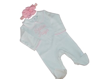 May include: White baby onesie with a pink bow headband. The onesie has a pink embroidered collar and the name "Hallie Paige" embroidered on the front.
