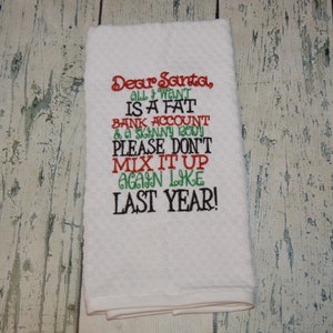 May include: White kitchen towel with red, green, and black embroidered text that reads "Dear Santa, All I want is a fat bank account & a skinny body. Please don't mix it up again like last year!"