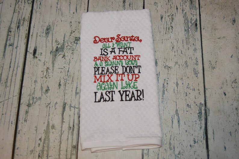 May include: White kitchen towel with red, green, and black embroidered text that reads "Dear Santa, All I want is a fat bank account & a skinny body. Please don't mix it up again like last year!"