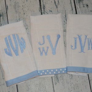 May include: Three white burp cloths with blue embroidered monograms. The monograms are "JW", "JW", and "JW". One burp cloth has a blue and white polka dot trim, one has a solid blue trim, and one has a solid blue trim.