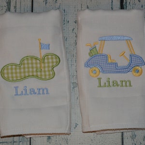 May include: Two white burp cloths with embroidered golf-themed designs. One cloth features a green golf course with a flag and the name "Liam". The other cloth features a blue golf cart with the name "Liam".