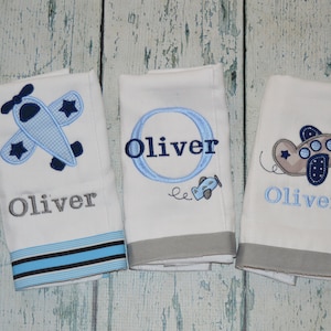 May include: Three white baby towels with airplane embroidery. Each towel features the name "Oliver" embroidered in gray or blue, with airplane designs in blue and gray. One towel has a blue and black striped border.