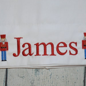 May include: White fabric with red embroidered nutcracker figures on either side of the name "James".