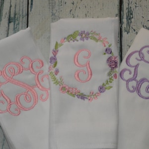 May include: Three white fabric burp cloths with embroidered monograms. The first cloth has a pink monogram with a floral wreath. The second cloth has a pink monogram with a single letter G in the center. The third cloth has a purple monogram.