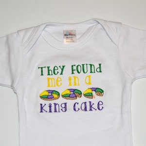 May include: White baby onesie with the text "They found me in a king cake" embroidered in green, yellow, and purple. Three purple, green, and yellow king cake donuts are embroidered below the text.