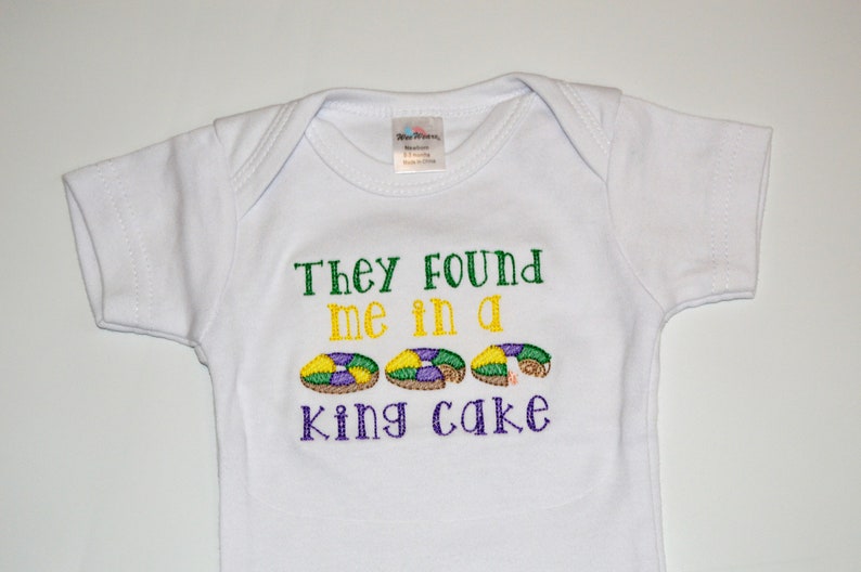 May include: White baby onesie with the text "They found me in a king cake" embroidered in green, yellow, and purple. Three purple, green, and yellow king cake donuts are embroidered below the text.