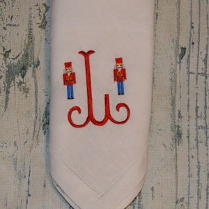 May include: White linen napkin with red embroidered nutcracker figures and a red embroidered letter "J" in the center.